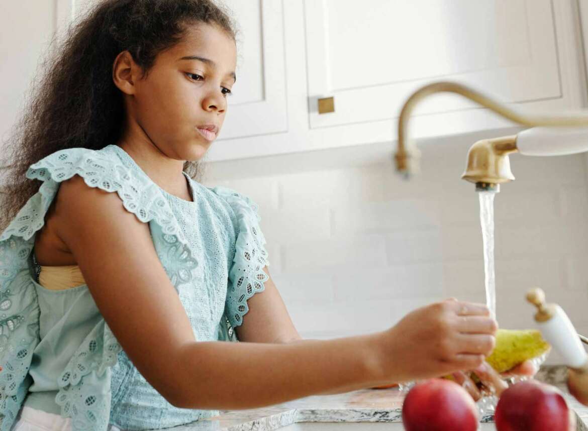 A young girl is washing fruits under running faucet water